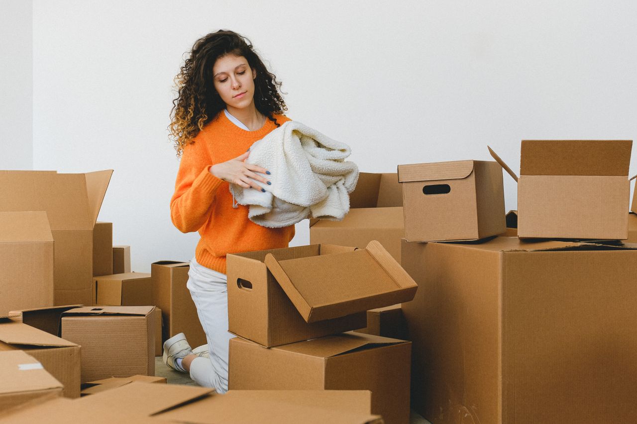 Services-02 Concentrated young lady with curly hair standing on knees on floor and packing clothes into carton boxes before moving into new flat